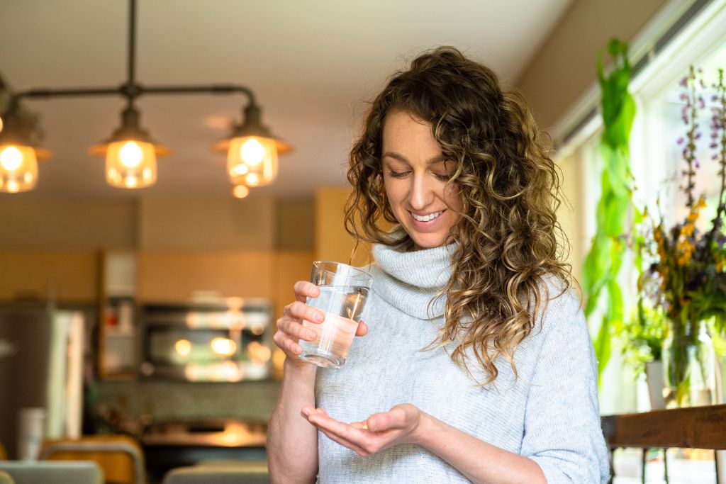 a woman holding a glass of water and looking at vitamin in her hand.