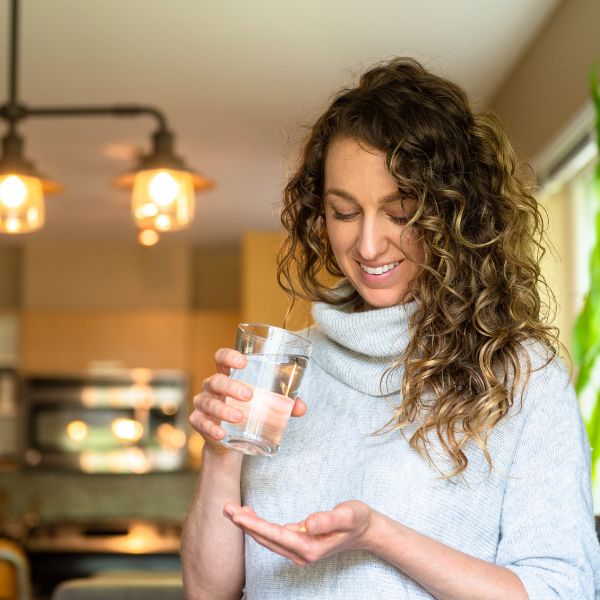 a smiling woman holding her supplements in her hand and a glass of water in the other hand.